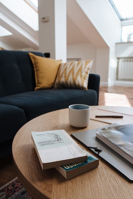 A woman wearing glasses and a grey and black t-shirt is using a handheld vacuum cleaner to clean a sofa armrest in a living room. The room features a tiled floor, a grey fabric sofa with plush cushion